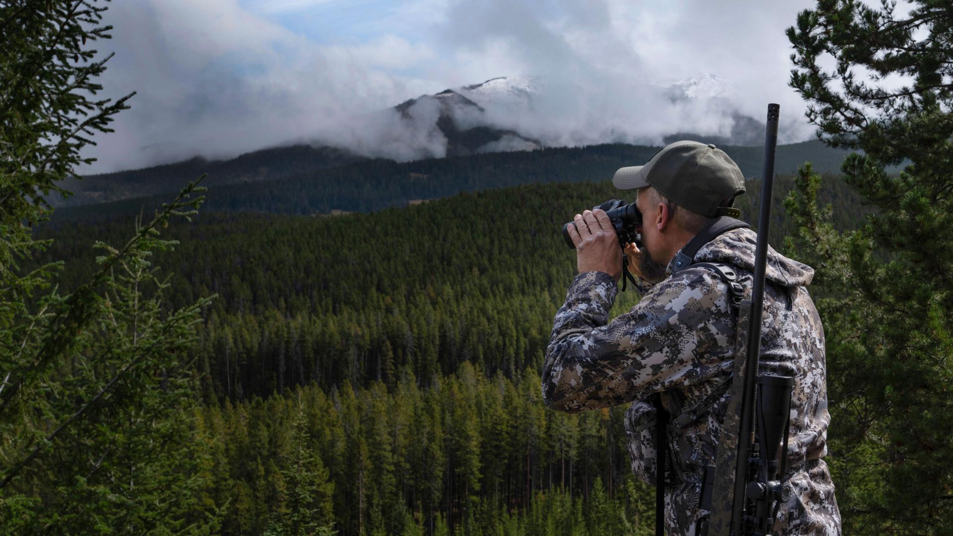 A hunter in camouflage gear uses ZEISS binoculars to scan a dense forest landscape with mountains and clouds in the background.