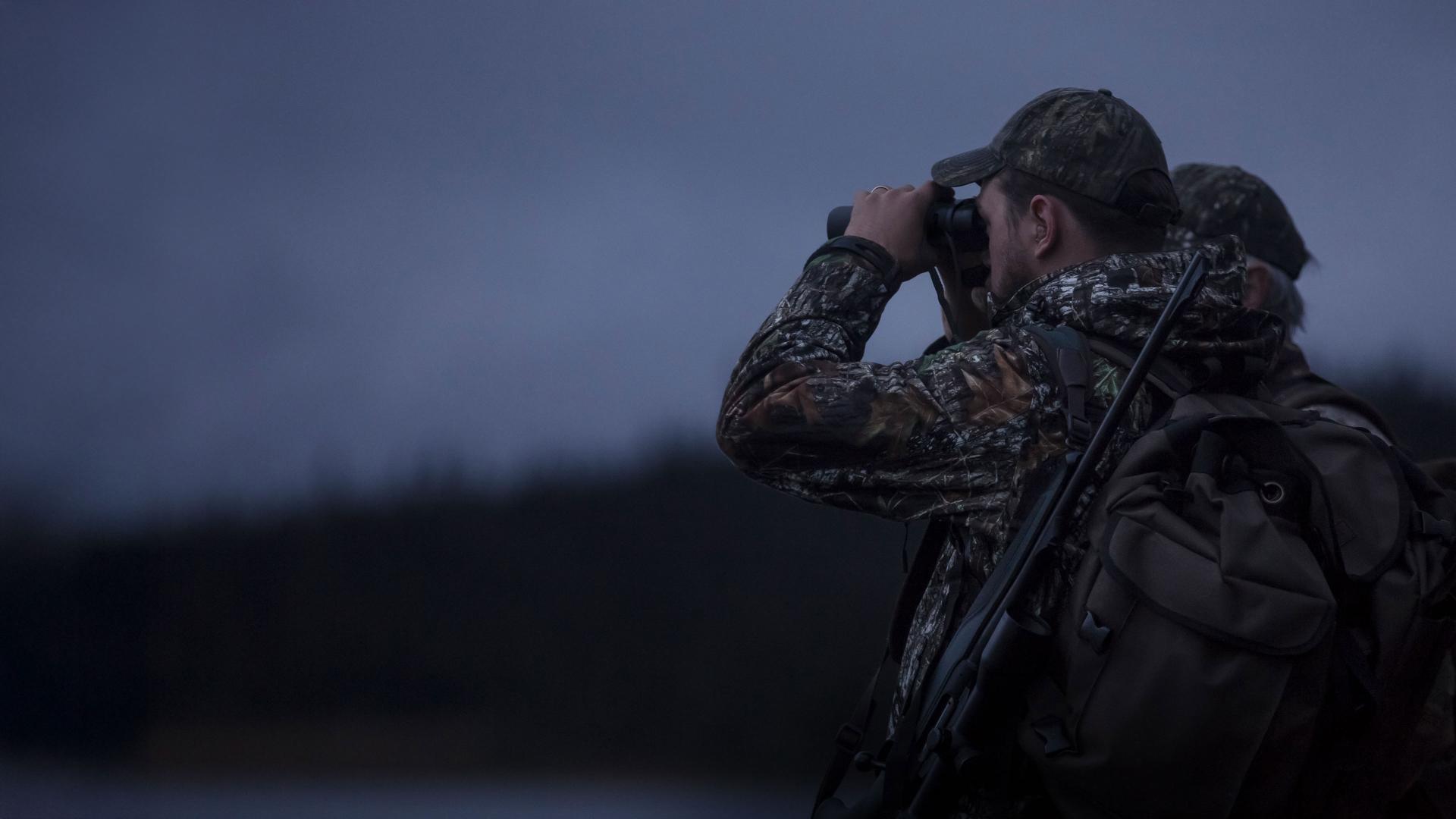 Hunter in camouflage gear using ZEISS binoculars at dusk with a forest background.
