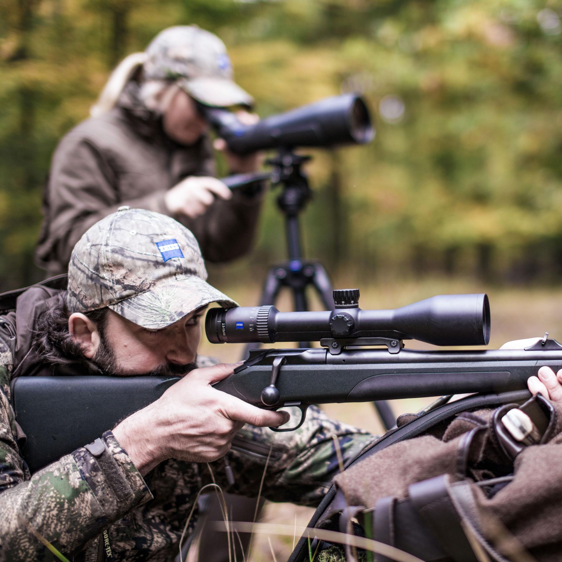 Hunter in camouflage aiming from a prone position with a rifle equipped with a ZEISS Conquest V6 2.5–15x56 riflescope, optimized for long-range accuracy and low-light conditions.