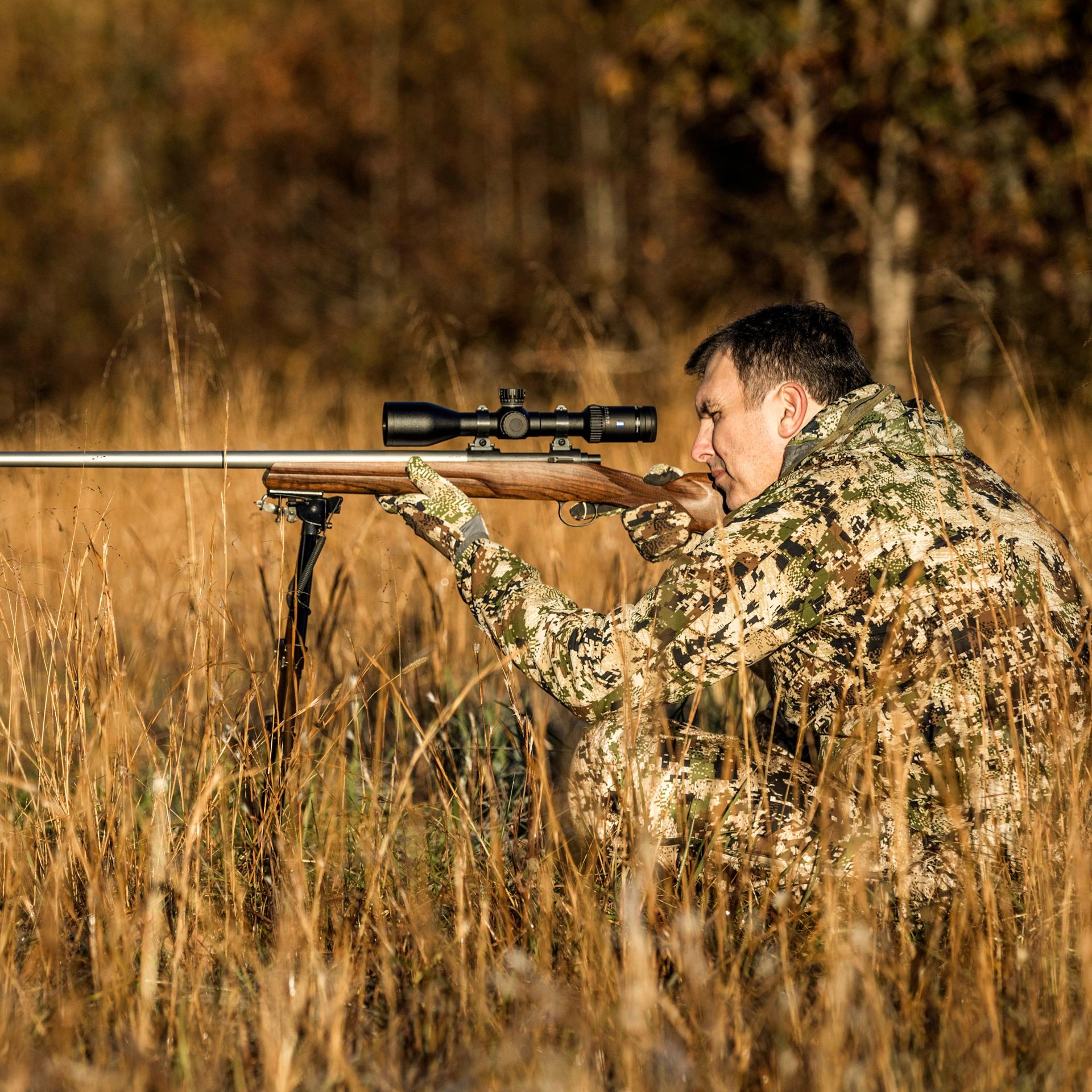 Hunter in camouflage clothing aiming across an open field with a rifle fitted with a ZEISS Conquest V6 3–18x50 riflescope, designed for versatile medium to long-range hunting.