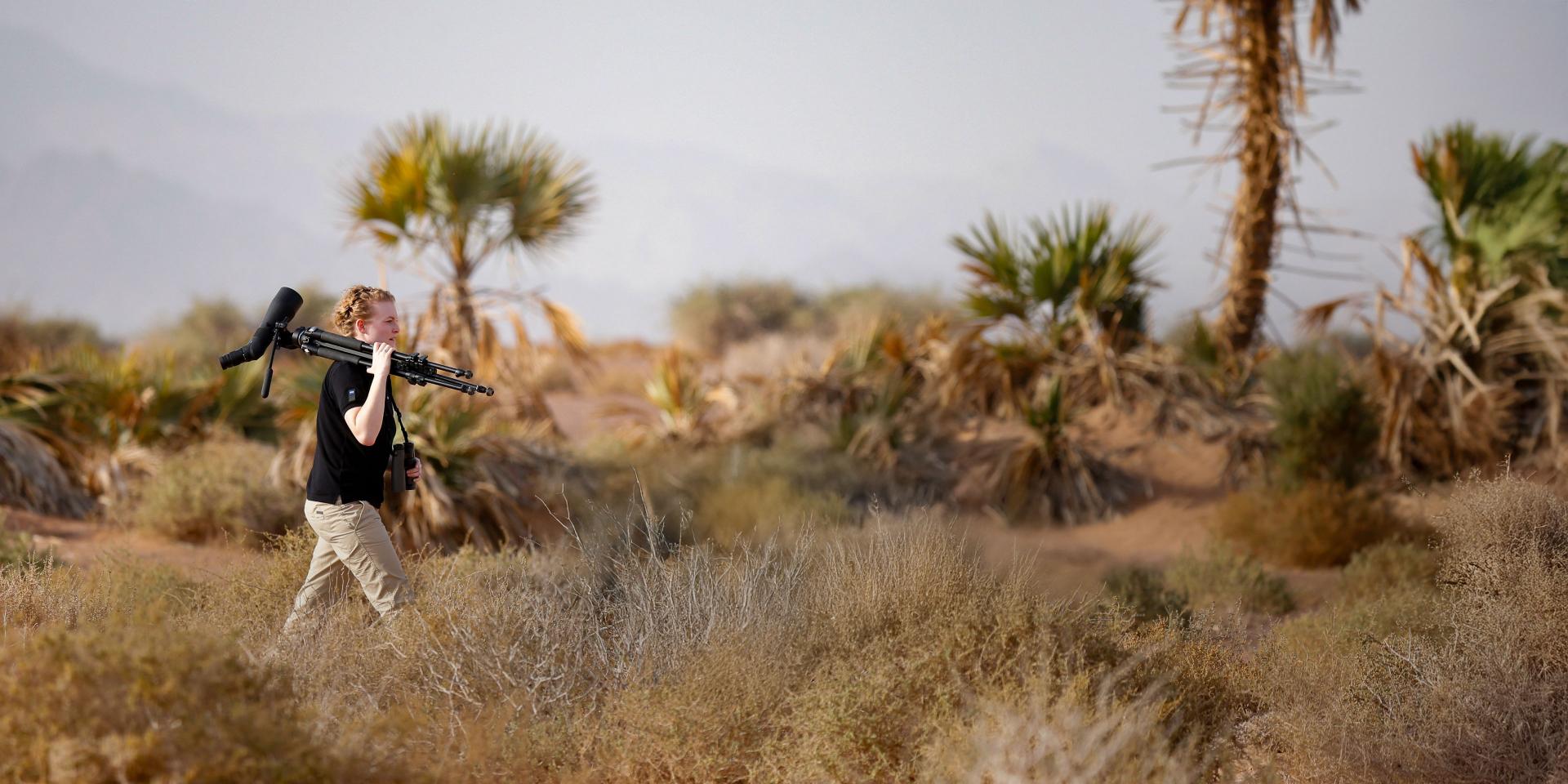 A person walks through a dry, desert-like landscape carrying a tripod and ZEISS spotting scope over their shoulder.