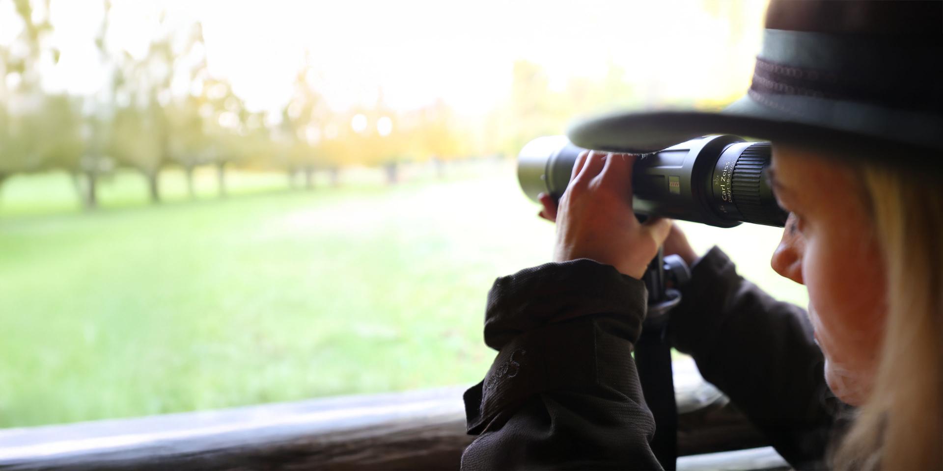 Person in outdoor clothing looking through a ZEISS binocular while observing a green field and trees.