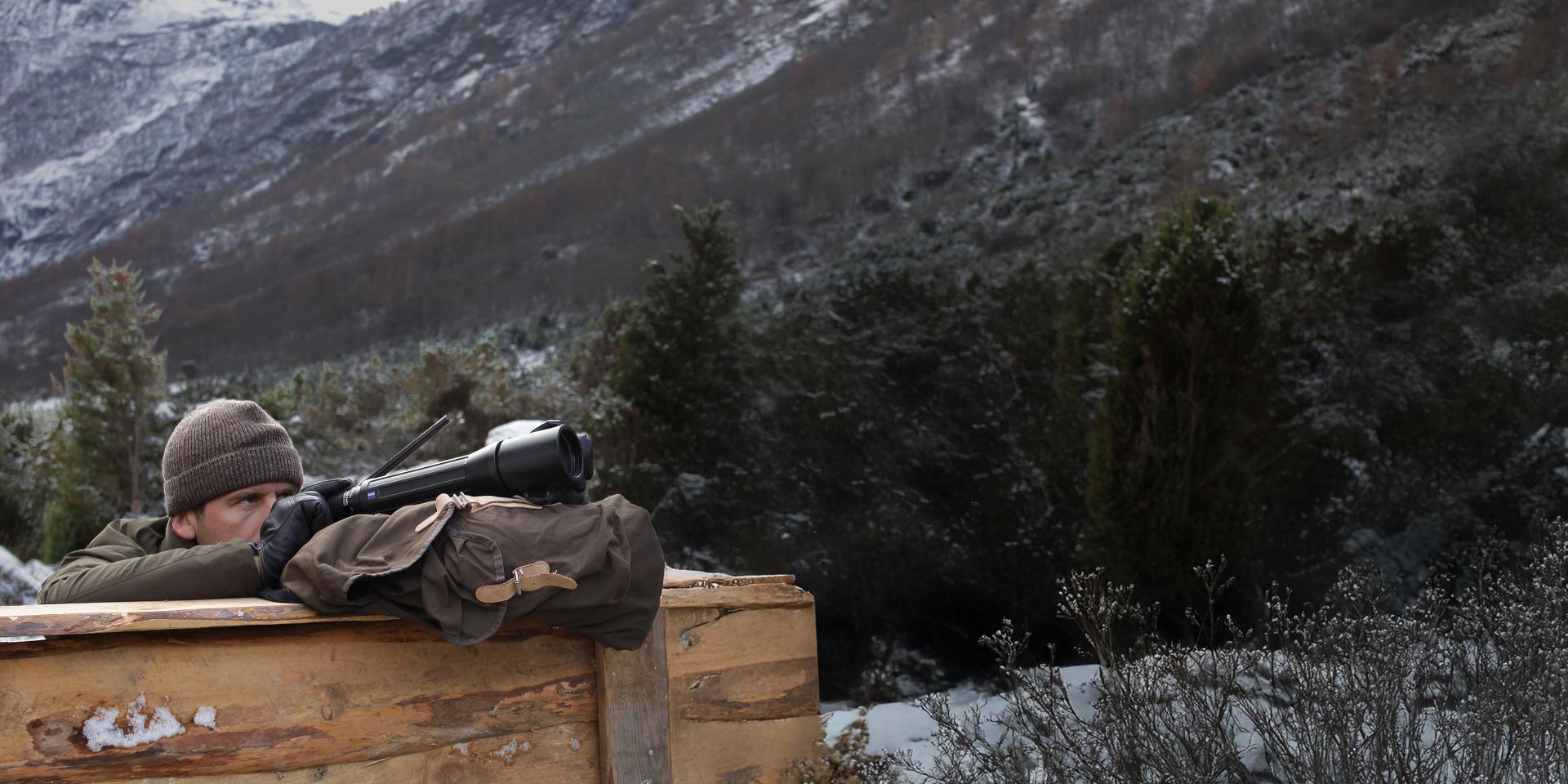 A person in winter clothing uses a ZEISS spotting scope while positioned behind a wooden barrier in a snowy mountain landscape.