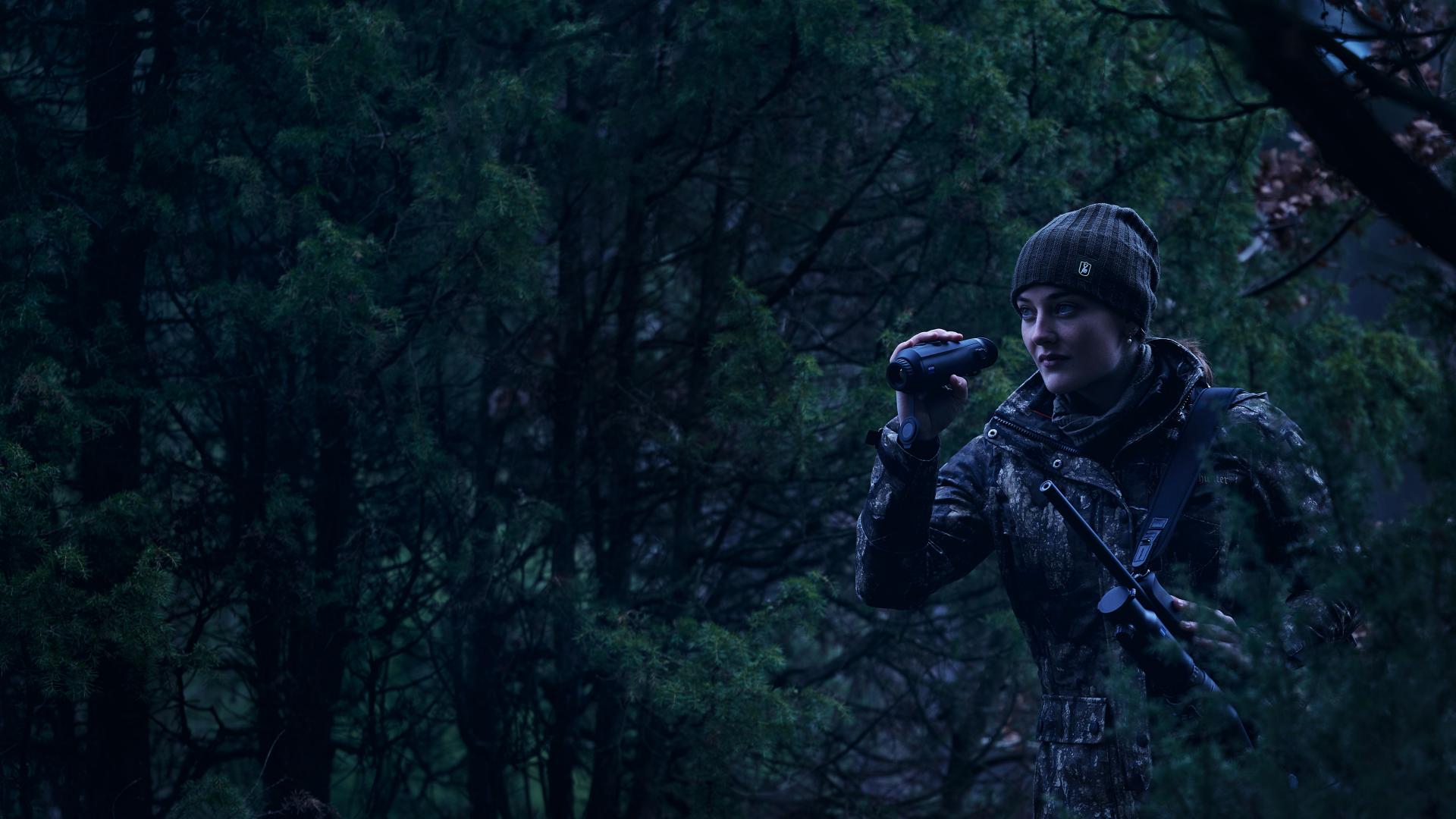 Person in camouflage gear using a ZEISS optic device while standing in a dense forest at dusk.