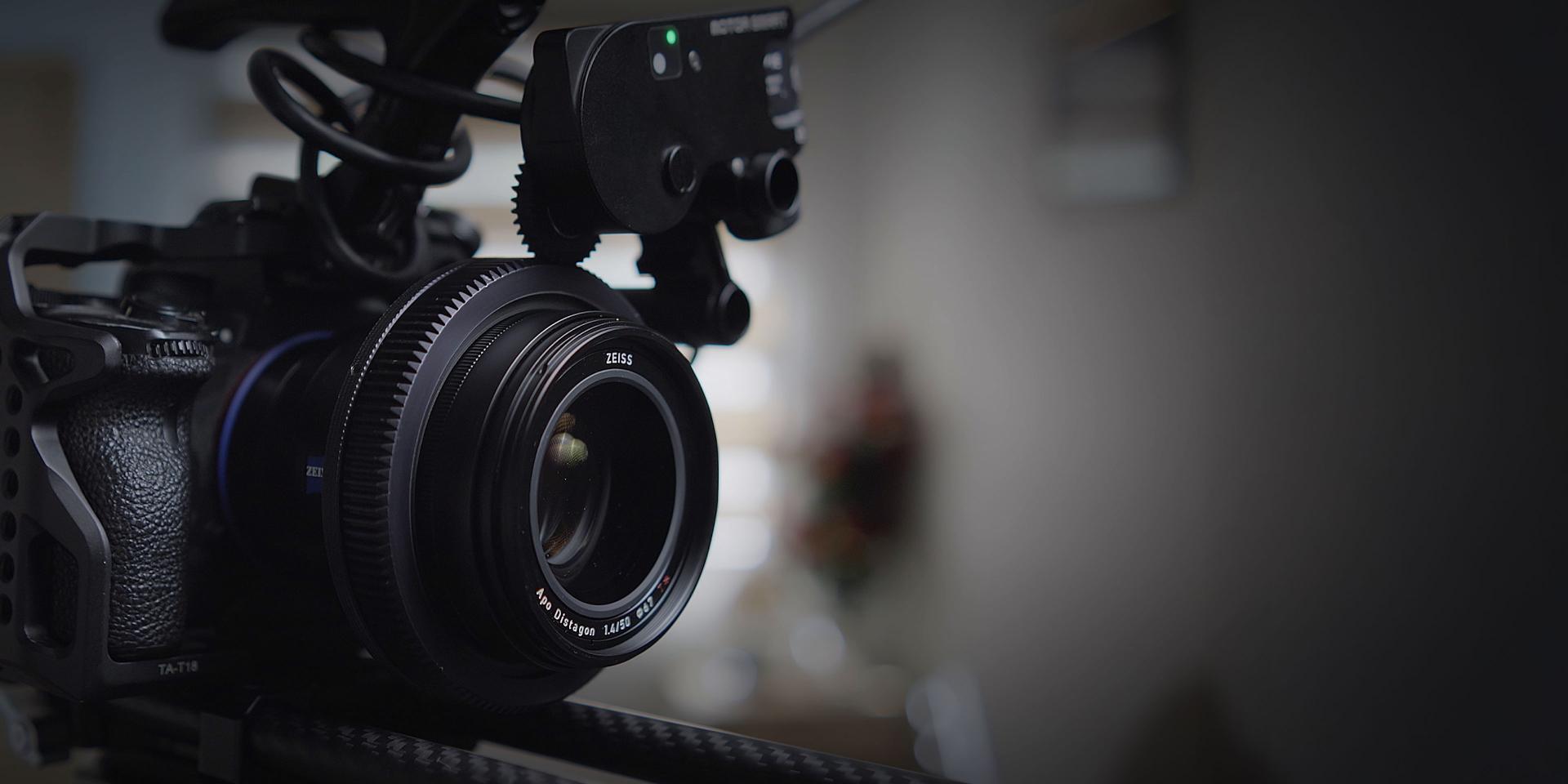 A videographer kneels on outdoor steps filming a person standing in white sneakers using a professional camera rig, with strong sunlight casting shadows.