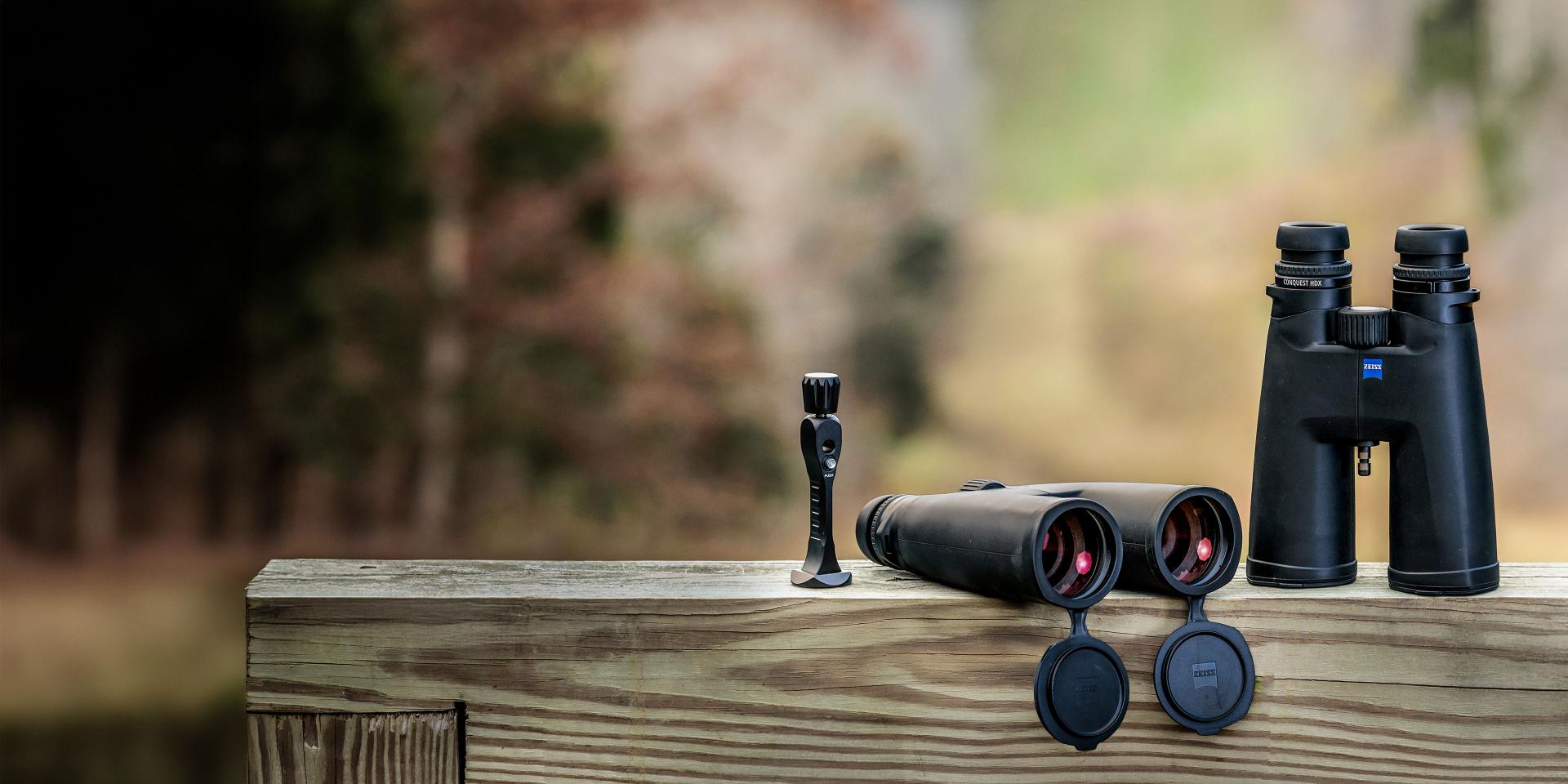 Three ZEISS optical devices, including a riflescope, a pair of binoculars lying flat with lens caps open, and a standing pair of binoculars, are displayed on a wooden railing with a blurred natural background.