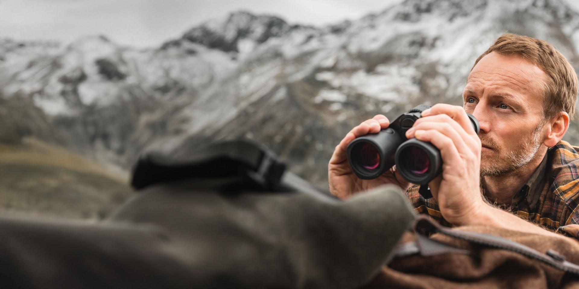 A man outdoors looks through ZEISS binoculars with a mountainous landscape in the background.