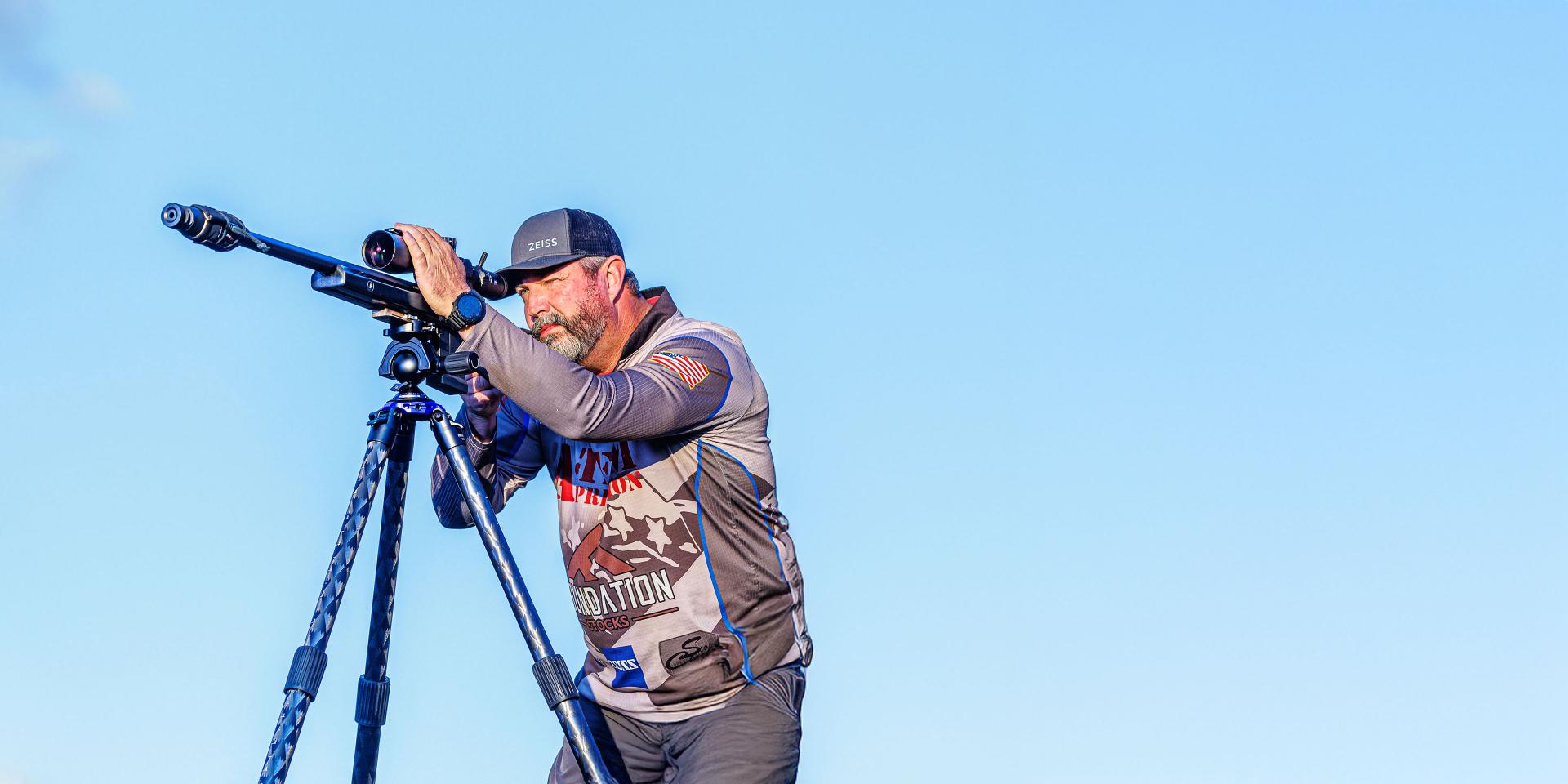 A man is using ZEISS optics mounted on a tripod while aiming outdoors under a clear blue sky.