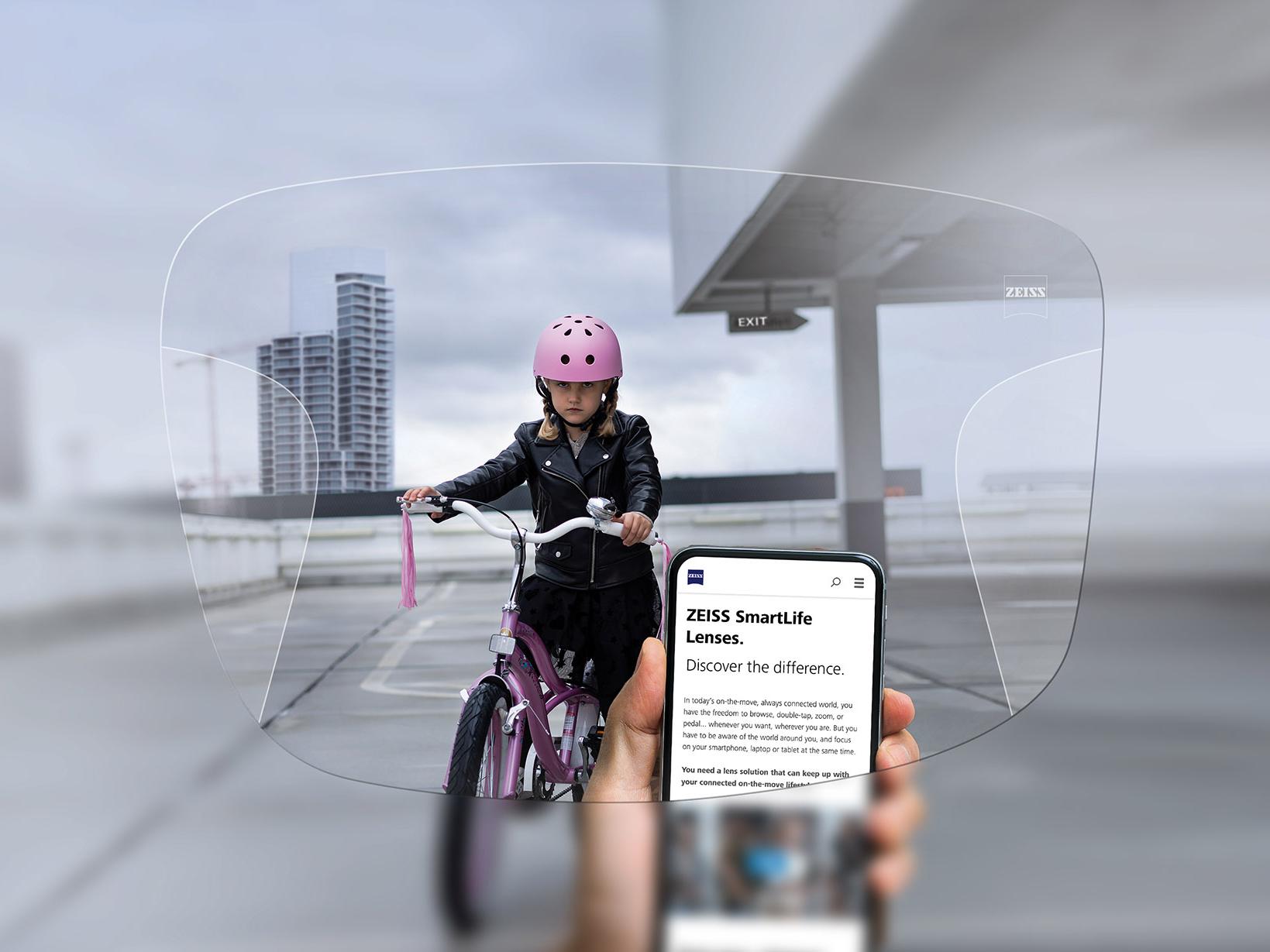 ZEISSSmartLife 遠近両用レンズでスマホを見る人。背景にピンクのヘルメットを被って自転車に乗る女の子が見える。 