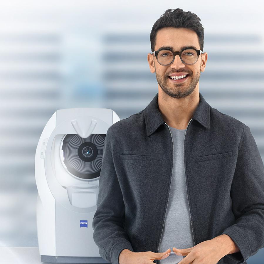 ZEISSのレンズを着けて、カメラに向かって微笑む茶色い短髪の若い男性。背後にZEISS i.Profiler plusがある。