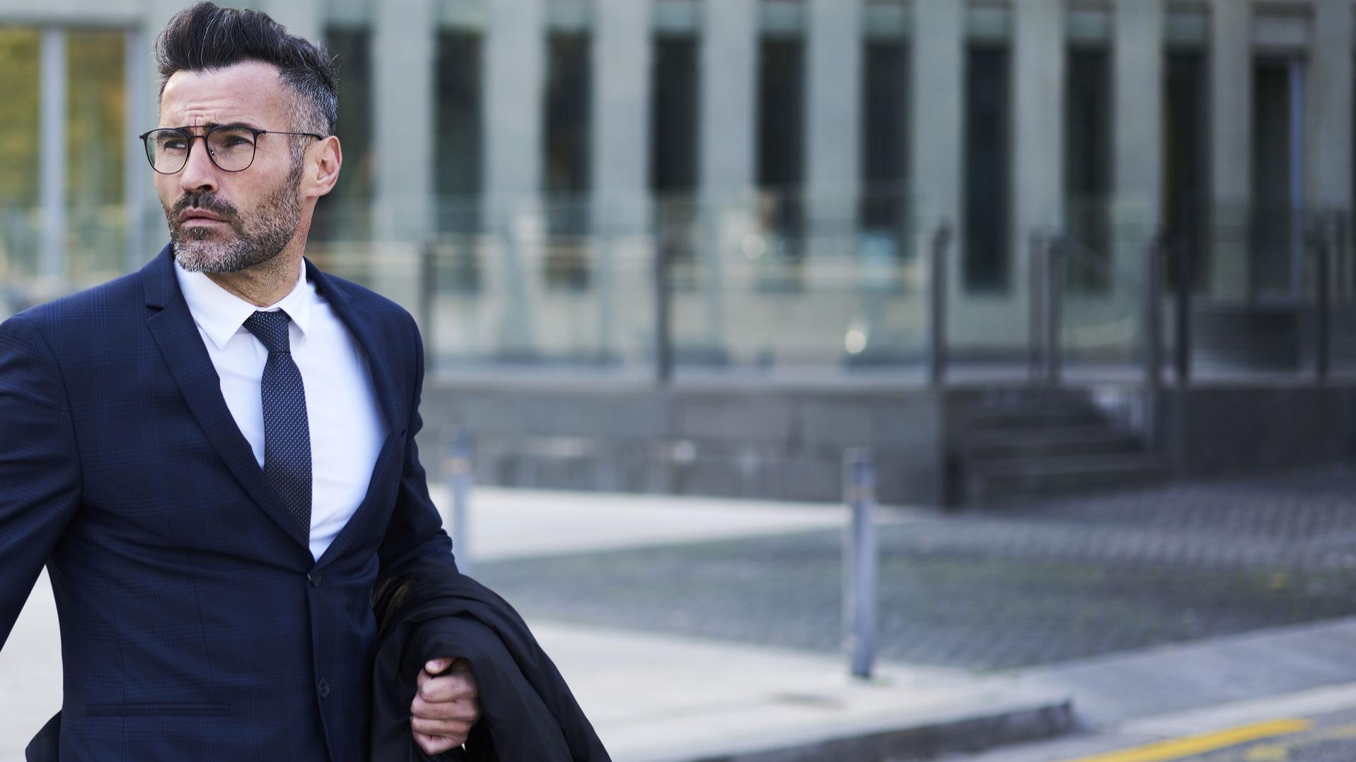 confident handsome bearded male lawyer in trendy eyeglasses crossing street passing modern constructed architecture buildings with headquarters in business district of city hurrying for meeting.