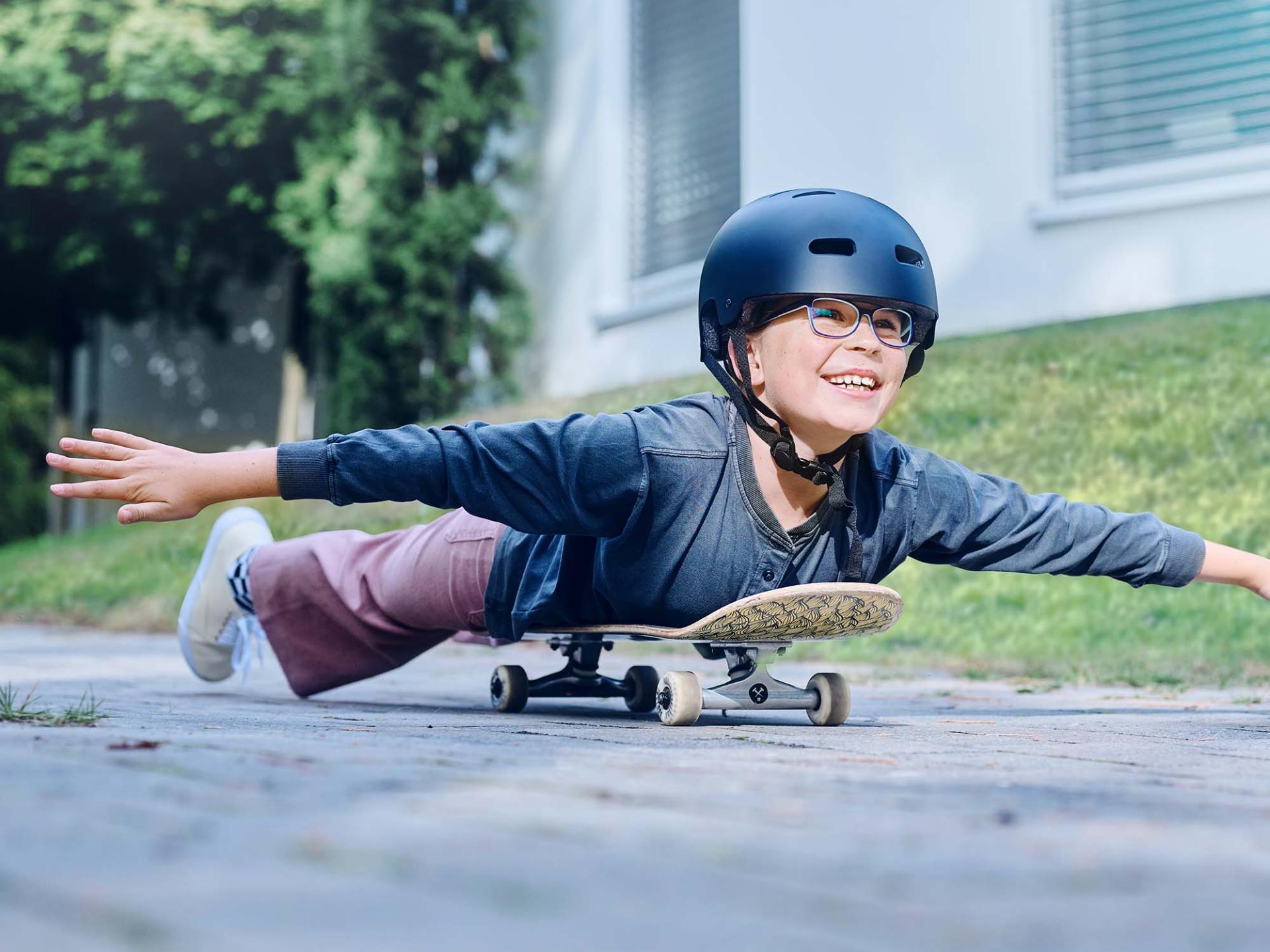 ヘルメットとメガネを付け、スケートボードに寝そべり、両手を広げて坂道を下る少女。